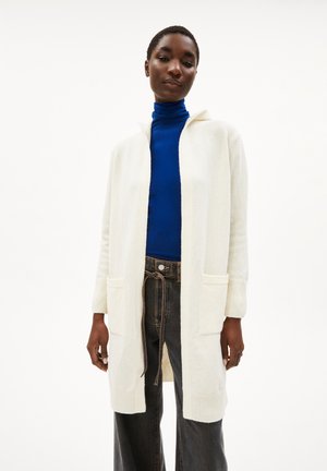 Witte gebreide cardigan met een capuchon, voorzien van twee voorzakken en geribbelde manchetten. Gekocht over een blauwe coltrui, gecombineerd met donkere jeans.