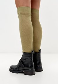 Medias de muslo de canalé en verde oliva, combinadas con botas de cuero negras hasta el tobillo que cuentan con detalles de hebilla y un tacón grueso.