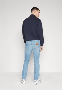 Felpa navy con colletto alto abbinata a jeans azzurri chiari con una toppa in pelle. Modello che indossa sneakers bianche, mostrando un look casual.