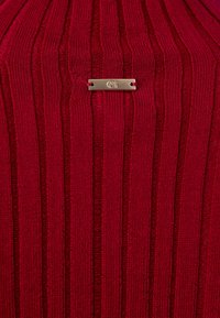 Tissu tricoté côtelé rouge avec une petite étiquette rectangulaire dorée gravée "CH" attachée au centre.