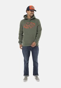 Hoodie vert olive avec logo orange, poche avant et capuche à cordon. Associé à un jean bleu et des chaussures à damier noir et blanc.