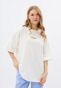 Overgrote witte katoenen t-shirt met korte mouwen en groene tekst die "MIRONS" op de voorkant leest. Gecombineerd met lichtblauwe gestreepte broek.
