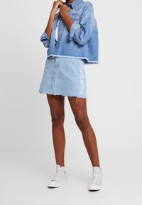 Osoba nosící světlou džínovou bundu a sladěnou sukni s bílou květinovou výšivkou, doplněné bílými vysokými teniskami.