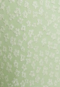 Tecido verde claro com uma superfície texturizada, apresentando padrões florais bordados em branco, espaçados uniformemente por todo o material.