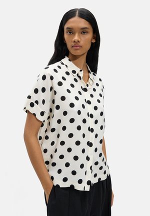 Kobieta o długich czarnych włosach, ubrana w białą krótką bluzkę z dużymi czarnymi kropkami i czarne spodnie, stoi z rękami w kieszeniach.