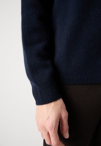 Sötétkék kötött pulóver bordázott mandzsettával és szegélyezéssel, amely puha textúrát mutat; sötét, testhezálló nadrággal viselve. Kéz látható az ujjnál.
