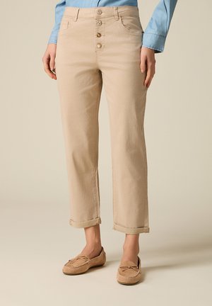 Beżowe spodnie z wysokim stanem i zapięciem na guziki, z podwiniętymi nogawkami, zestawione z beżowymi mokasynami oraz jasnoniebieską, długorękawową koszulą.