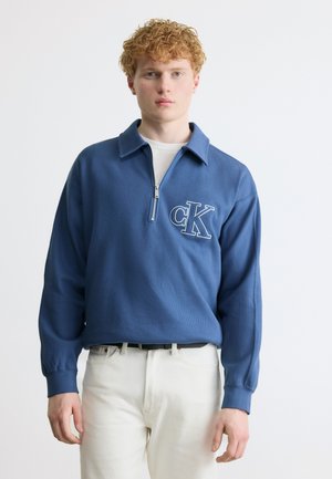 Młody mężczyzna z kręconymi rudymi włosami, ubrany w niebieski sweter zapinany na zamek do ćwiartki z logo "cK" oraz białe spodnie, na tle jasnego, jednolitego tła.