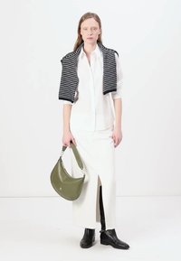 Jupe longue blanche avec une fente à l'avant, superposée à une chemise blanche à boutons et un pull noir à rayures drapé sur les épaules. Sac à main vert olive.