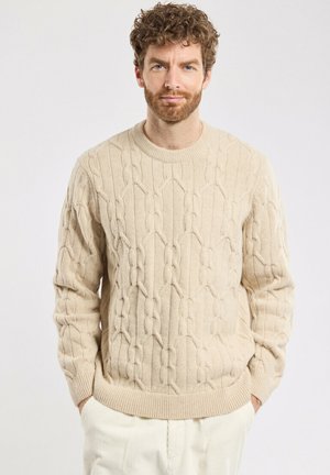 Beżowy sweter w splot kabelkowy z okrągłym dekoltem i prążkowanymi mankietami, z teksturowanym wzorem na całej długości. Noszony z jasnymi spodniami.