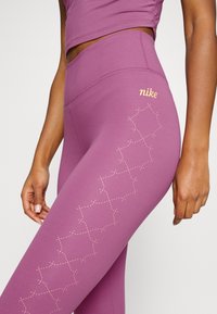 Paarse leggings gemaakt van een gladde, rekbare stof, met een gouden "Nike"-logo en een diamanten patroon in lichte stiksel langs de zijkant.