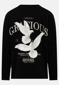 Fekete, hosszú ujjú póló fehér galambokkal és "KIRÁLYI" felirattal. A grafikák között vonalkód mintázat és szövegelemek találhatók. Puha pamut anyag.