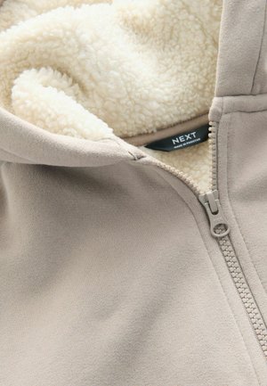 Beige zip-up hoodie med en mjuk krämfärgad fleecefoder. Har en framdragskedja och etikett. Materialet verkar slätt och mysigt.