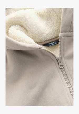 Beige mikina na zip s měkkou krémovou fleecovou podšívkou. Má přední zip a štítek. Materiál vypadá hladce a útulně.