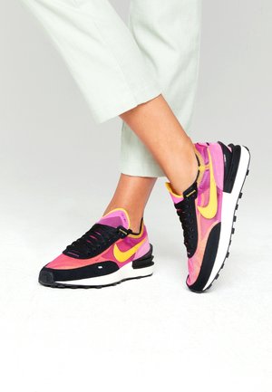 Personne portant un pantalon blanc et des baskets Nike aux couleurs vives rose et jaune, debout sur un sol blanc.