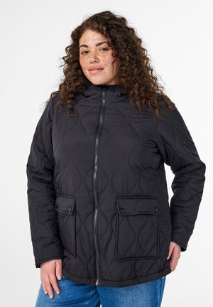 Lockige Person mit Sommersprossen, die eine schwarze gesteppte Jacke mit Vordertaschen und blaue Jeans trägt, steht vor weißem Hintergrund.