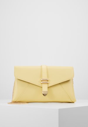 Keltainen kirjekuorimallinen clutch-laukku, jossa on kultainen ketjuhihna, keskellä pystysuora hihna ja kullanvärinen solki valkoisella jalustalla.