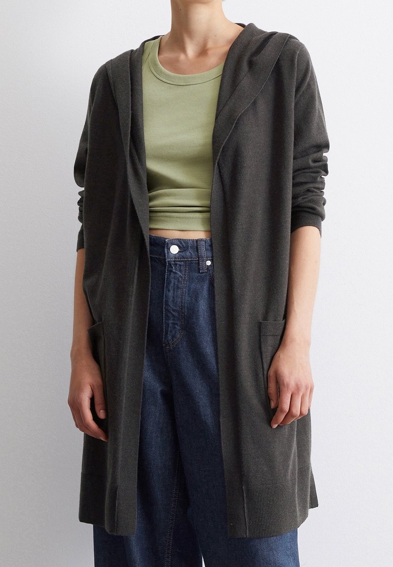 Dark gray knit cardigan with a hood, long sleeves, and front pockets. Worn over a light green ribbed tank top and high-waisted blue jeans.