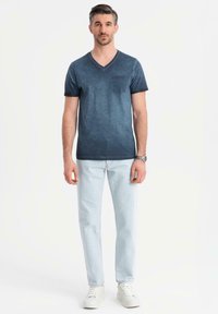 Tmavě modré tričko s výstřihem do V a malou náprsní kapsou, nošené s světle modrými džíny a bílými teniskami. Bavlněná tkanina s lehkou strukturou.