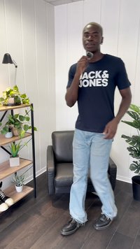 Crna majica s bijelim natpisom "JACK & JONES", svijetloplave traperice, crne cipele, stojeći u sobi s biljkama i crnim naslonjačem.