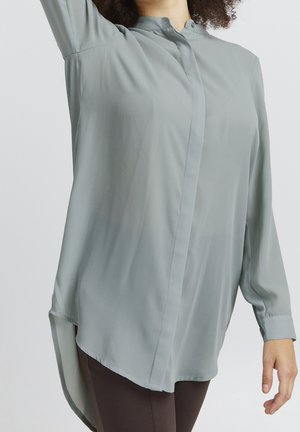 Camicia tunica verde chiaro realizzata in materiale trasparente. Presenta una chiusura con bottoni frontale, maniche lunghe e un orlo posteriore più lungo con spacco sui lati.