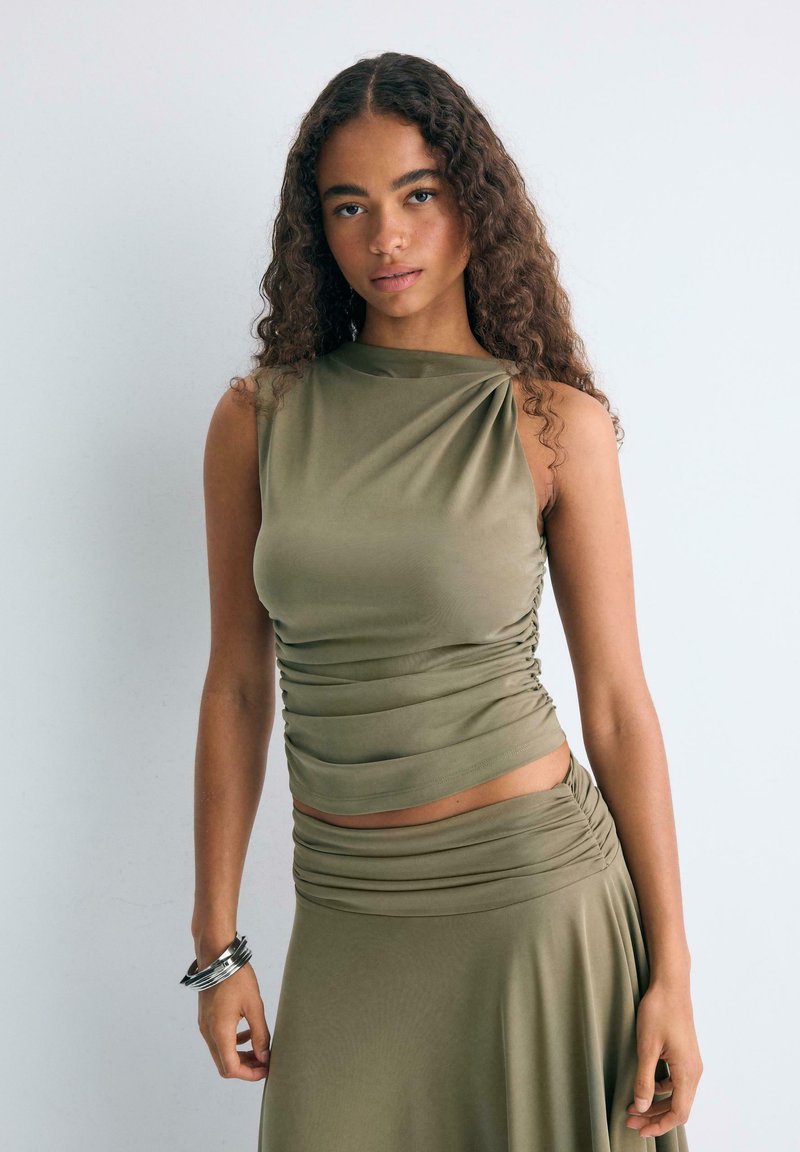 Jeune femme aux longs cheveux bouclés, portant un haut olive sans manches froncé et une jupe assortie, debout devant un fond blanc uni.