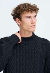 Pull en laine bleu marine foncé à col montant, avec un motif en câble texturé. Manches retroussées et design épais et chaud.