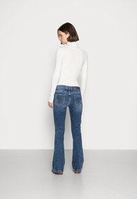 Flared blue denim jeans with light fading, stitched back pockets, and a white ribbed turtleneck. The model faces away from the camera.
