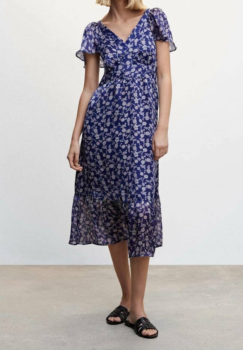 Robe florale bleue avec des manches courtes et volantes, design portefeuille, longueur mi-mollet, tissu transparent, et ourlet volé, associée à des sandales noires.