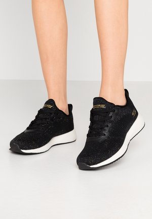 Benen met zwarte Skechers sneakers met witte zolen, staand op een lichtgrijze vloer tegen een effen achtergrond.