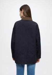 Nadmierna tmavomodrá bunda s hnedým golierom, vyrobená z hladkej látky; má zapínanie na gombíky na manžetách a rovný strih pre pohodlné nosenie.