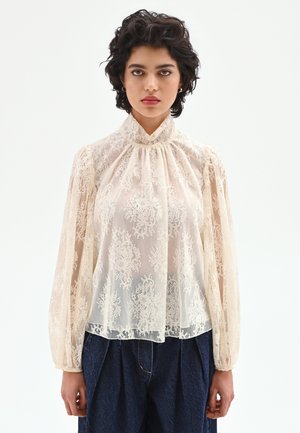 Blusa in pizzo color crema chiaro con maniche lunghe a sbuffo e collo alto. Caratterizzata da motivi floreali intricati e una texture morbida e trasparente.