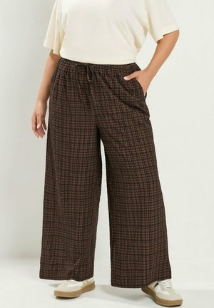 Persona con pantalones anchos de cuadros marrón y negro con cintura elástica ajustable y zapatillas blancas, de pie con una mano en el bolsillo.