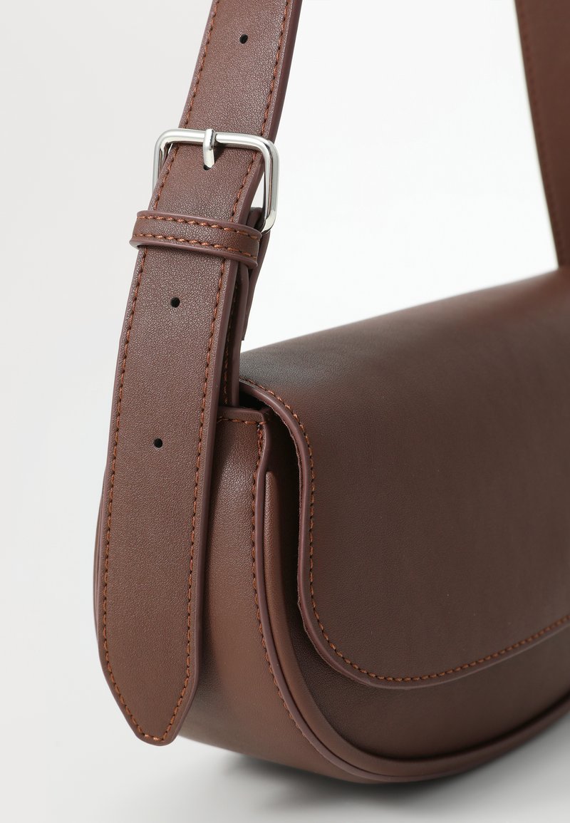 Bolso cruzado de cuero marrón con forma curva, laterales acolchados y correa ajustable con herrajes plateados y costuras en contraste.