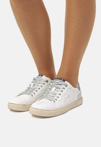 Zapatillas blancas con cordones plateados, paneles laterales texturizados y un acento gris en el talón. Suela de goma, diseño casual.