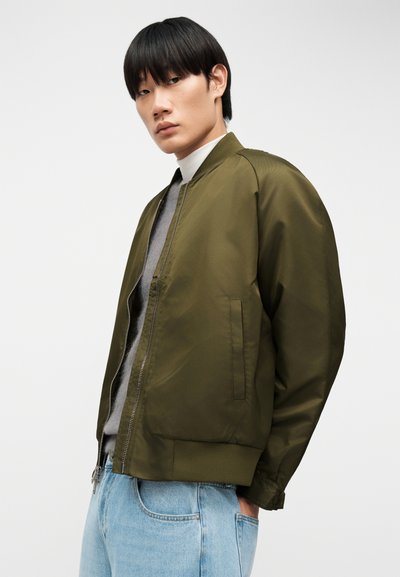 Νεαρός άνδρας που φοράει μπουφάν bomber ελιάς, γκρι πουλόβερ, λευκό μπλουζάκι με ψηλό γιακά και ανοιχτόχρωμο μπλε τζιν, στέκεται μπροστά σε απλό λευκό φόντο.