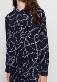 Chemise bleu marine à manches longues boutonnée avec motif chaînes blanches portée par une personne aux longs cheveux bruns sur fond uni.