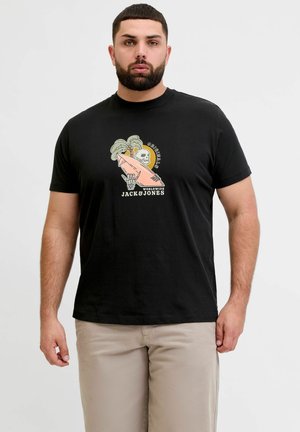 Άνδρας που φοράει μαύρο μπλουζάκι με γραφικό σκελετό που κάνει σερφ και φοινικόδενδρο, μπεζ παντελόνι, στέκεται σε απλό φόντο.