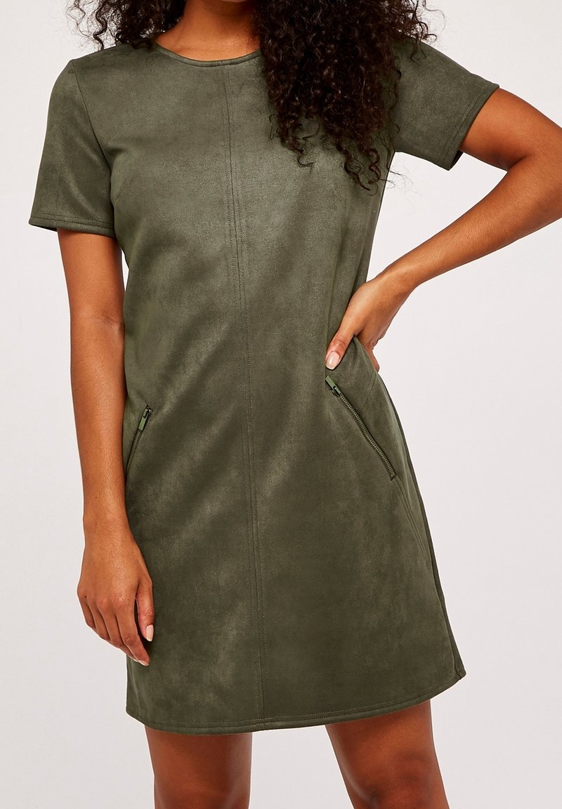 Femme portant une robe en daim vert olive à manches courtes avec des poches zippées à l'avant, main posée sur la hanche, debout devant un fond uni.