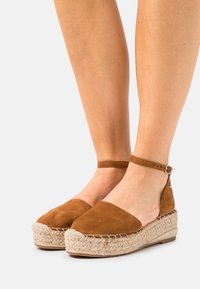 Brown suede espadrille wedges with a braided jute sole, open sides, and an adjustable ankle strap feature a rounded toe design.