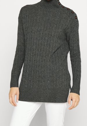 Mørkegrå kabelstrikket rullekravesweater med knapdetaljer på venstre skulder, båret med hvide bukser, hænder afslappet ved siderne.
