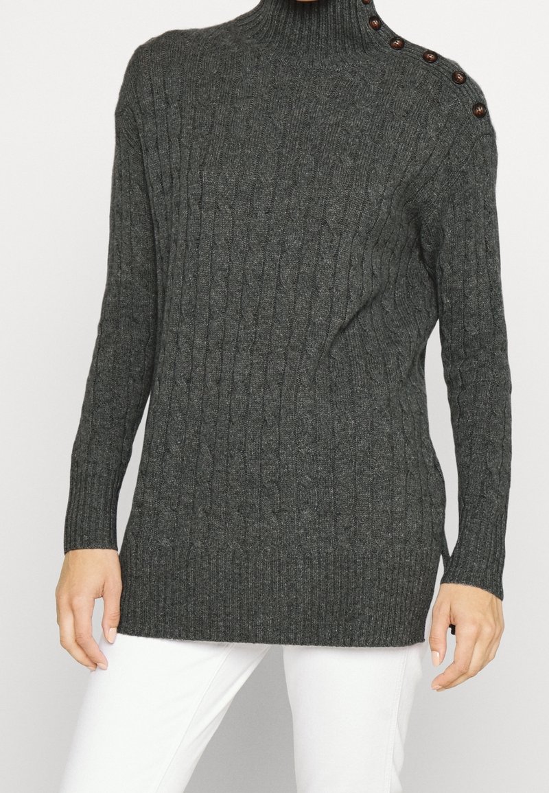 Pull col roulé gris foncé à maille torsadée avec des boutons sur l'épaule gauche, porté avec un pantalon blanc, mains détendues le long du corps.