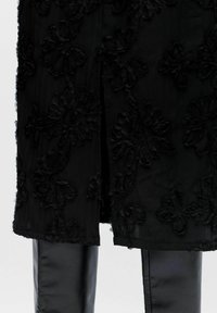 Sort gennemsigtig nederdel med tekstureret blomsterapplikation, delt foran, båret over skinnende sorte læderbukser mod en hvid baggrund.