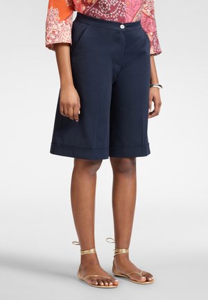 Donna che indossa pantaloncini sartoriali blu navy fino al ginocchio, una camicetta colorata a maniche lunghe, sandali piatti dorati con cinturino alla caviglia e un braccialetto d'argento.