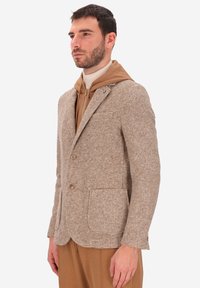 Uomo che indossa un blazer a quadri beige sopra un hoodie zip-up color sabbia e pantaloni coordinati color sabbia, rivolto leggermente a sinistra su uno sfondo bianco.