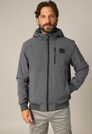 Veste zippée grise avec une section supérieure matelassée, capuche, deux poches avant et poignets élastiques. Comprend un petit patch logo sur la poitrine.