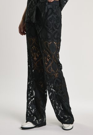 Pantalon large en dentelle noire avec des motifs floraux et géométriques complexes, porté avec des chaussures blanches à bouts pointus, visible de la taille jusqu'aux pieds.