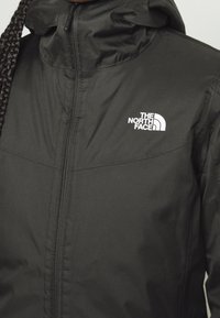 Veste noire The North Face avec fermeture éclair frontale, logo visible sur la poitrine, portée par une personne aux cheveux tressés.