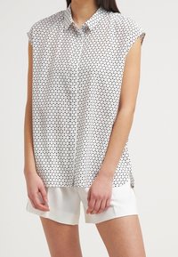 Ärmlös vit blus med svarta fyrkantiga prickar, matchad med enkla vita shorts på en stående person.