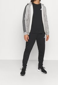 Sudadera gris con cremallera y rayas negras, camiseta negra y pantalones de chándal negros a juego. Zapatillas deportivas negras con acentos blancos.